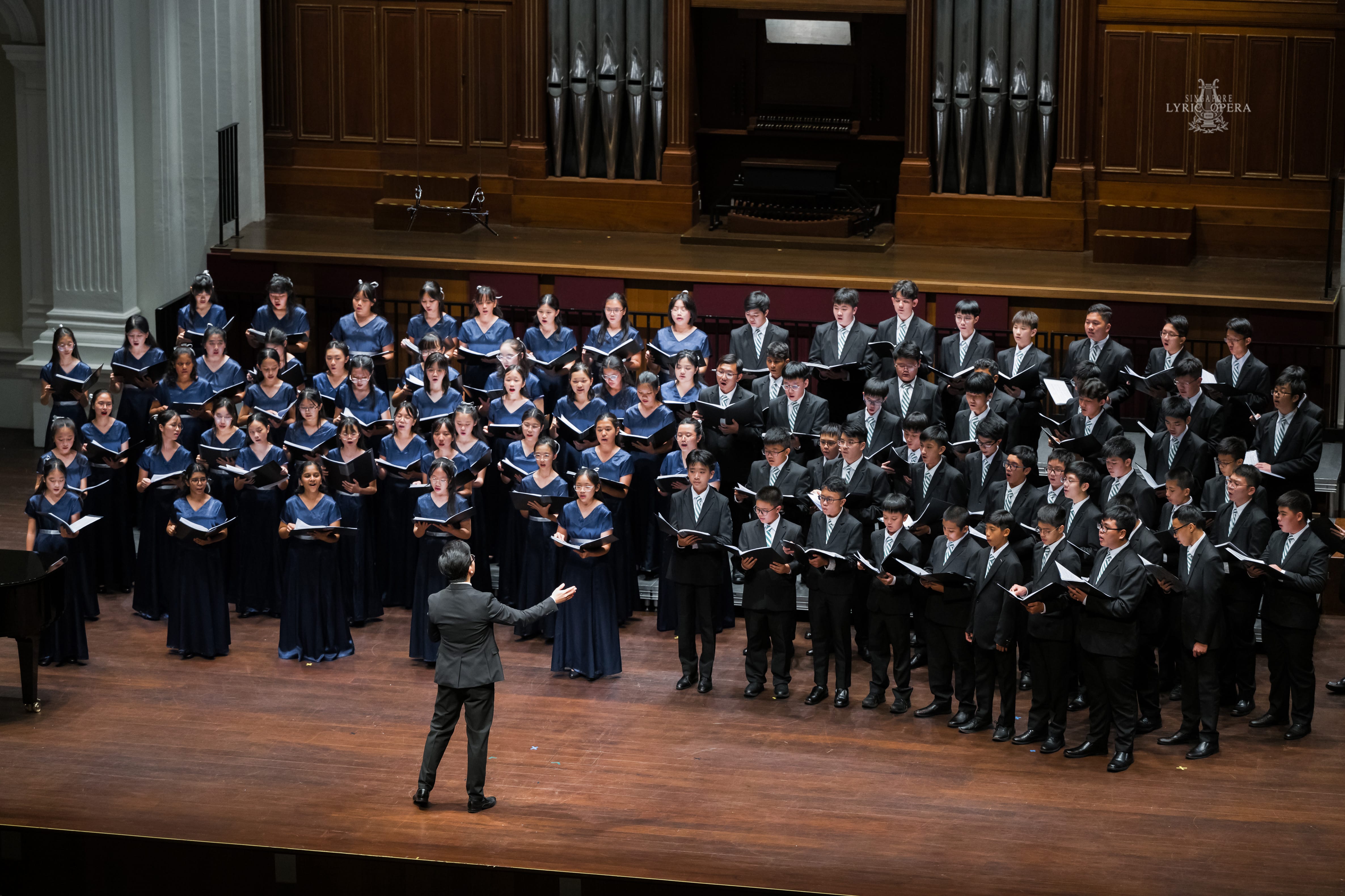 A conductor leads a large youth choir on stage, girls in blue gowns and boys in black suits holding score books.